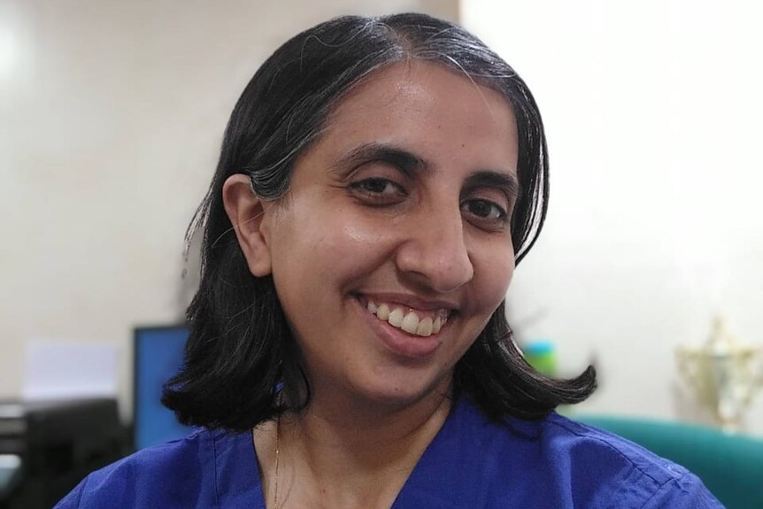 An Indian woman in blue scrubs smiles for the camera
