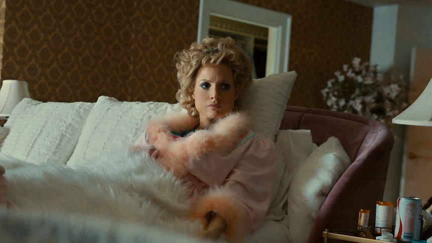 Middle-aged woman with curly blonde hair reclines on lush white sofa, wearing a soft, plushy pink dressing gown.