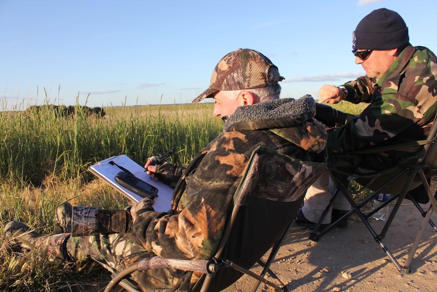Two men wearing camouflage suits sit outdoors in camping chairs, surrounded by high grass. One is holding a clipboard and pen.