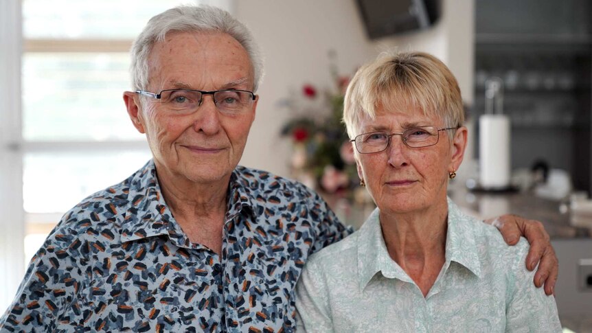 An older couple sits together. He has his arm around her shoulders. They smile gently at the camera.