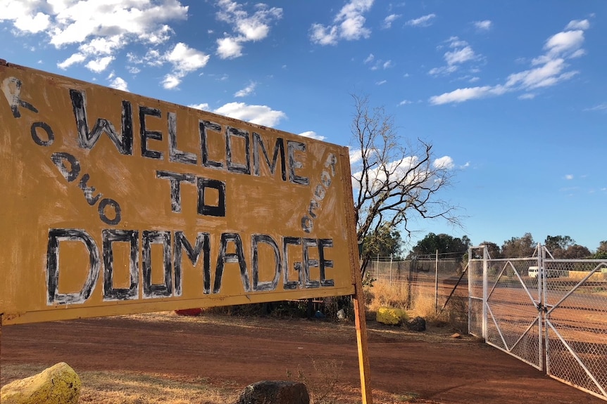 A faded sign in red dirt by the road reads 'Welcome to Doomadgee'.