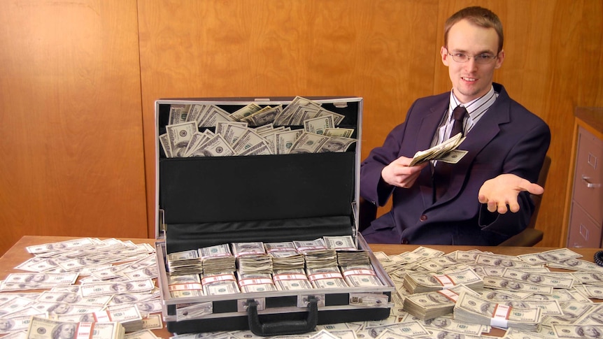 young man in a suit sits at a desk with piles of cash