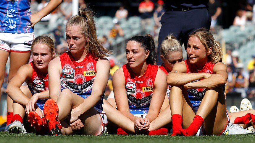 Eliza McNamara, Sinead Goldrick, Eden Zanker and Megan Fitzsimon of the Melbourne Demons AFLW team sit on the grass looking sad.