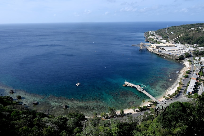 A view from a hill looking down over a tropical forest to a large bay with a jetty, yacht, housing and industrial area.