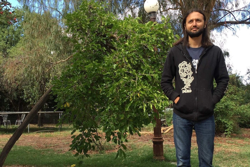 A man in his mid-30s man with long brown hair standing in front of trees and a lamp-post.