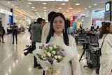 A young woman grins as she stands in a busy airport with a bunch of flowers.