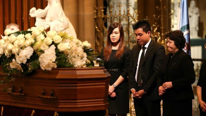 Three Asian Australians stand in front of a coffin covered in white flowers, the mother looking visibly emotional