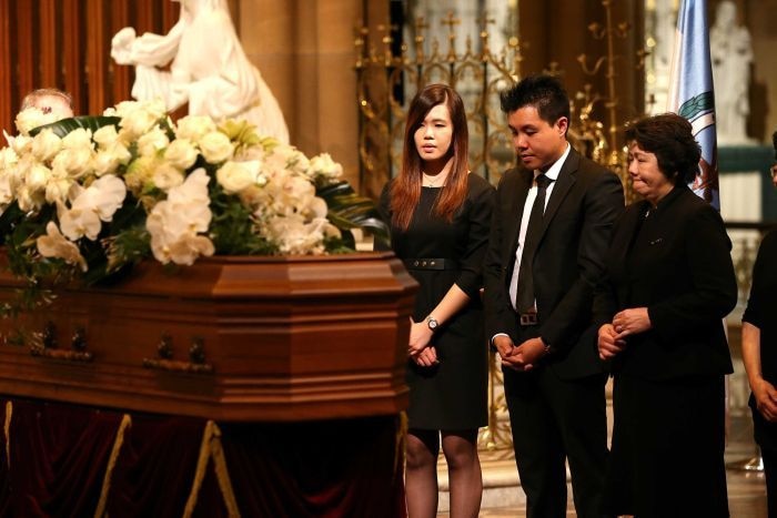 Three Asian Australians stand in front of a coffin covered in white flowers, the mother looking visibly emotional