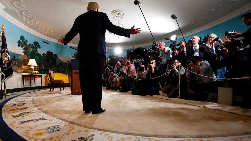 A view of the back of Donald Trump standing with his arms outstretched, facing dozens of reporters, photographers and TV cameras