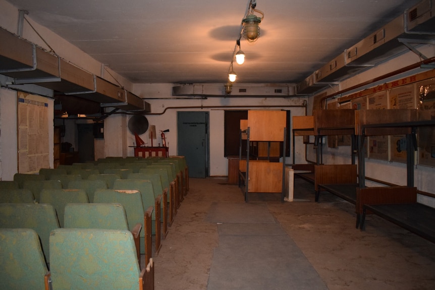 Rows of chairs are lined up on one side of a dark, concrete room, while empty bunkbeds line the other wall