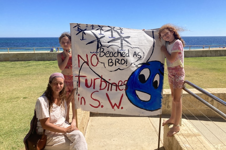 A woman and two girls hold up a handmade sign reading 'beached as bro! no turbines in S.W.'