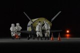 A group of four people in space suits inspect a small plane on a runway at night time.