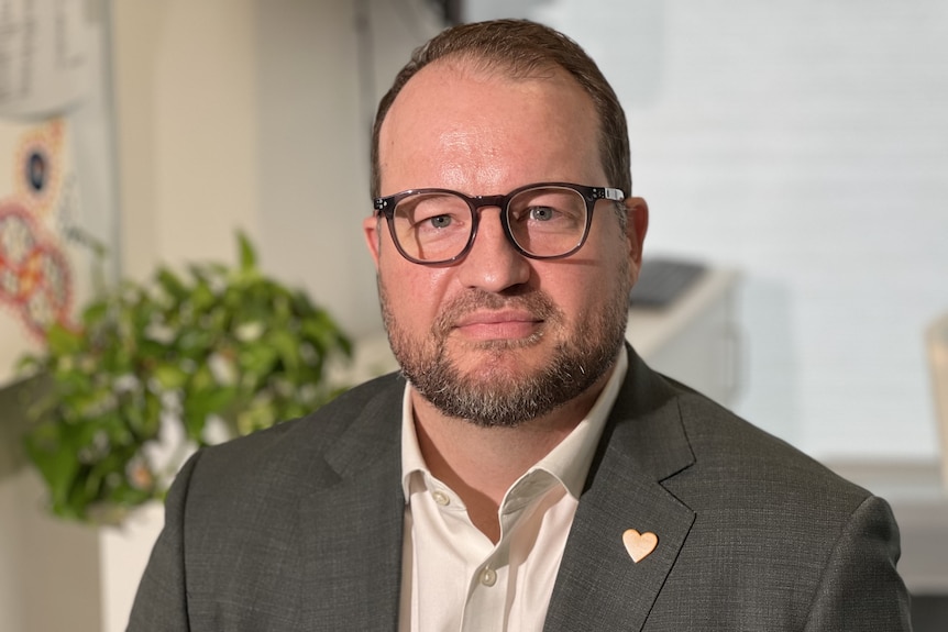 A white man with short brown hair and glasses. He is in an office wearing a grey suit