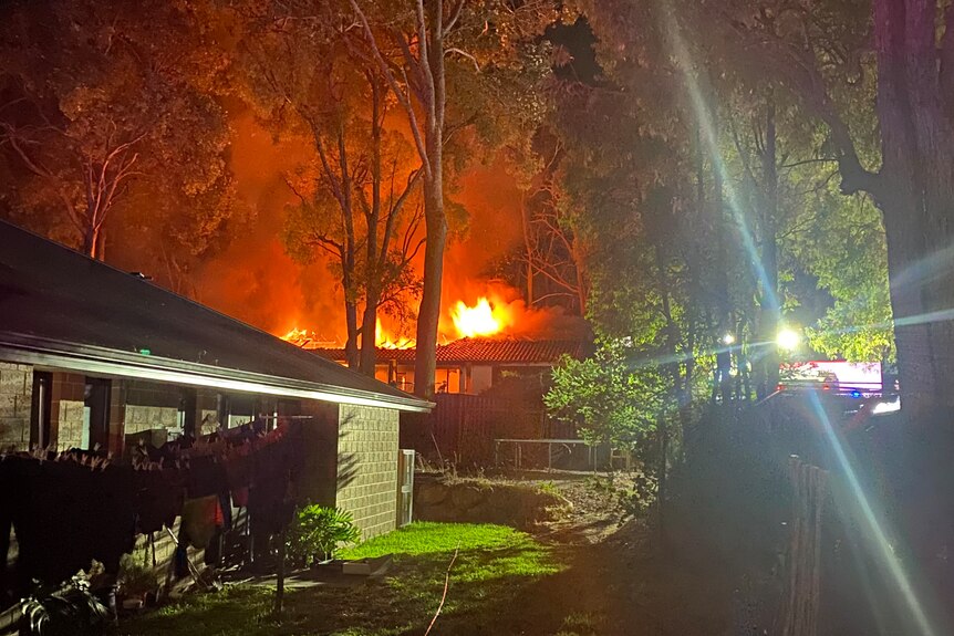 Flames rise up from a house at night, seen from the backyard of a neighbouring property.
