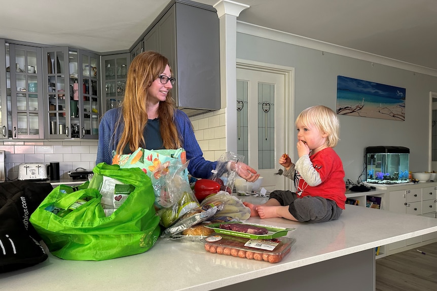 mother unpacking shopping in kitchen with toddler on bench