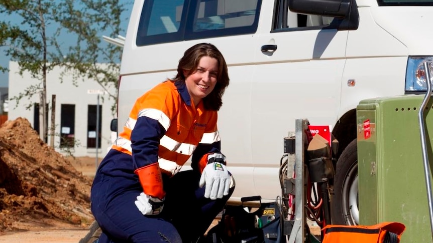 women working on electrical supply point kneels in front on her work and smiles at camera