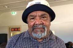 An aboriginal elder stands in a room wearing a white hat.