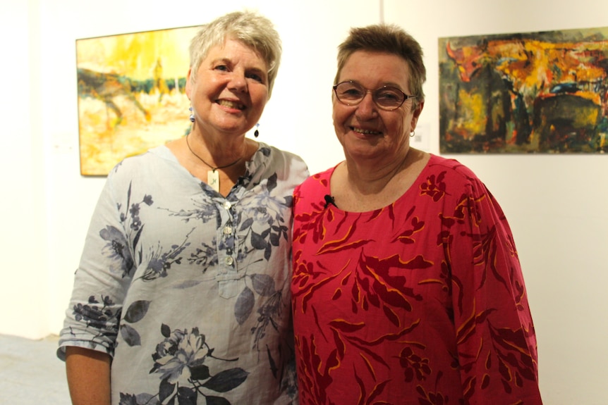 Two older women stand close, smile, both short-haired, one wears grey patterned blouse, other pink, art hanging behind.