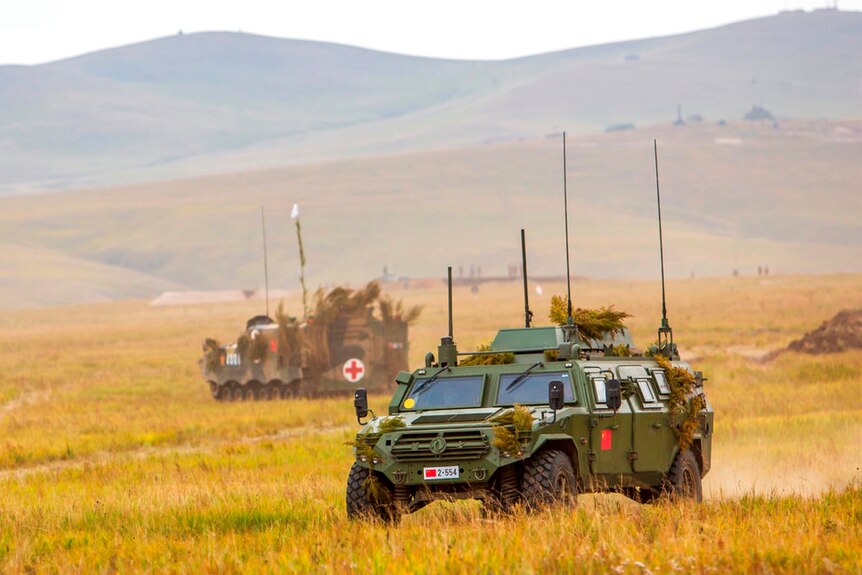 Two green-painted military vehicles with multiple antennae roll through a flat grassy area with hills in the background