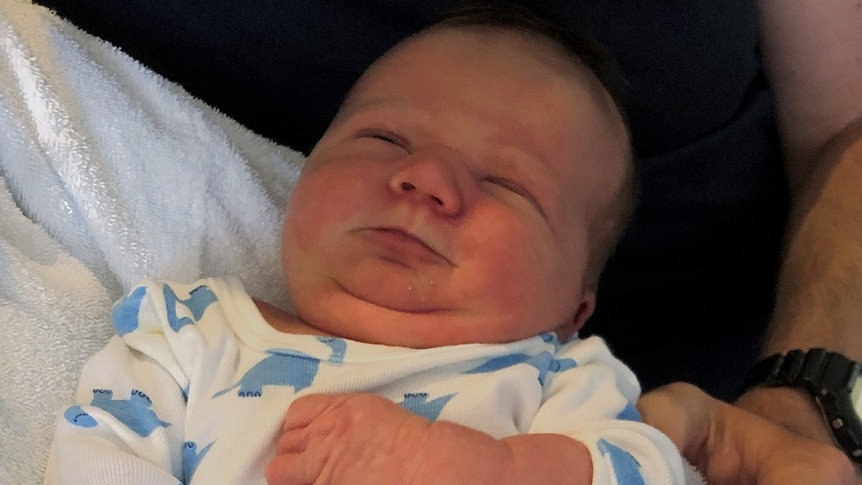 Newborn baby wearing white and blue one piece and grey socks suit being held in parents' arms smiles with eyes closed