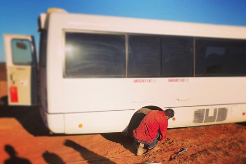 a white school bus stands on a red dirt road while a person changes its tyre