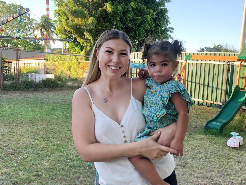 A young mother wearing a white shirt holds a toddler smiling at the camera