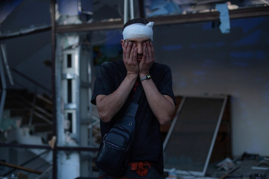 A man with bandage on his head covers his eyes and he stands outside damaged building.