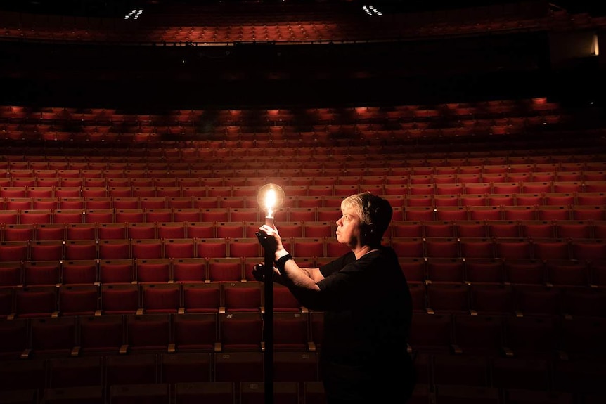 A woman holds a large light globe with a dark theatre behind her