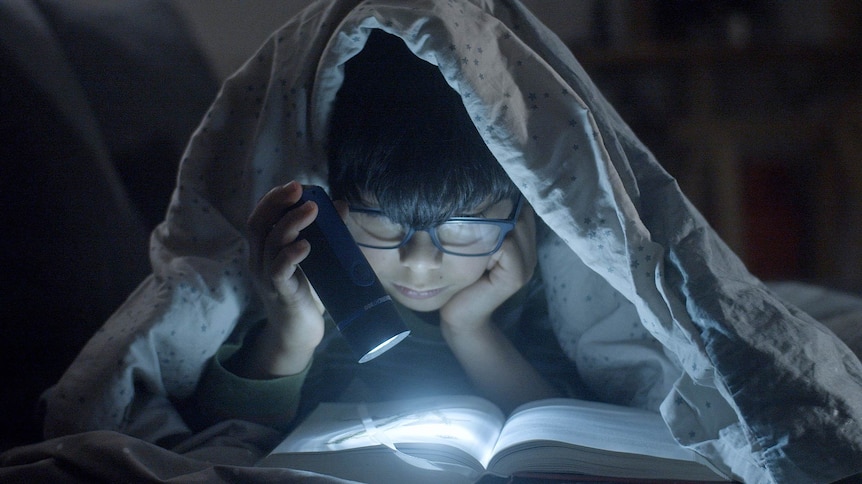 An Asian boy under his bed covers using a flashlight to read a thick book.