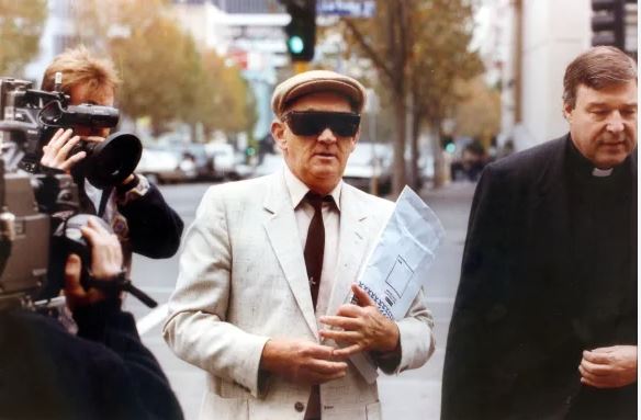 A man with a white cap and black sunglasses walks between a TV cameraman and another man dressed in black.