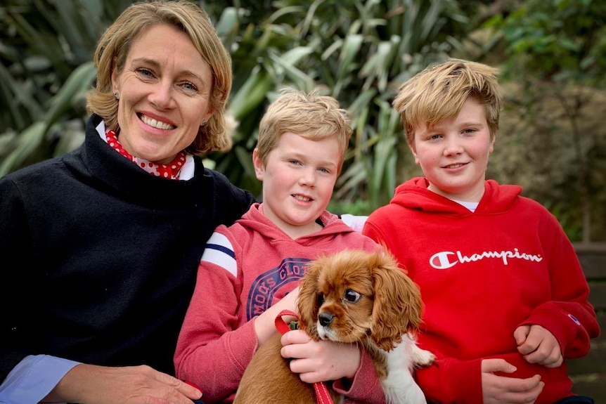 A woman with short blonde hair sits besides to boys on a garden bench. One boy holds a small orange and white dog on his lap.