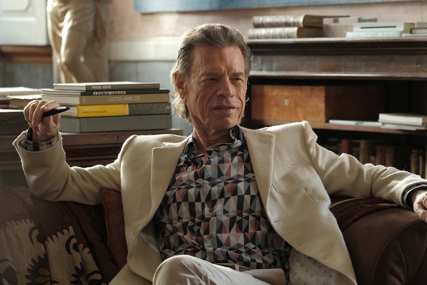 An older man in beige suit and patterned shirt sits on couch in daylight lit interior room with books and bookshelves.