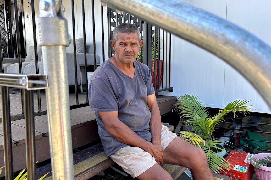 A First Nations man with grey hair sits on a stairs leading to a verandah, with a solemn expression on his face.