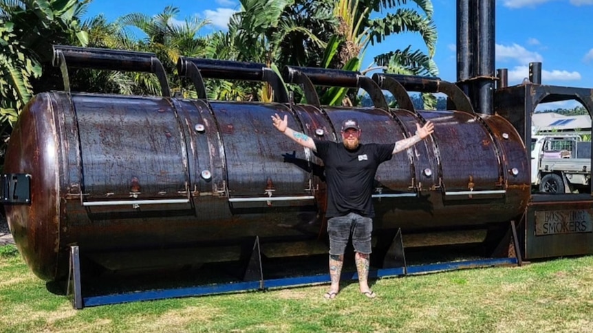 A man wearing black waves his arms in front of a giant brown meat smoker