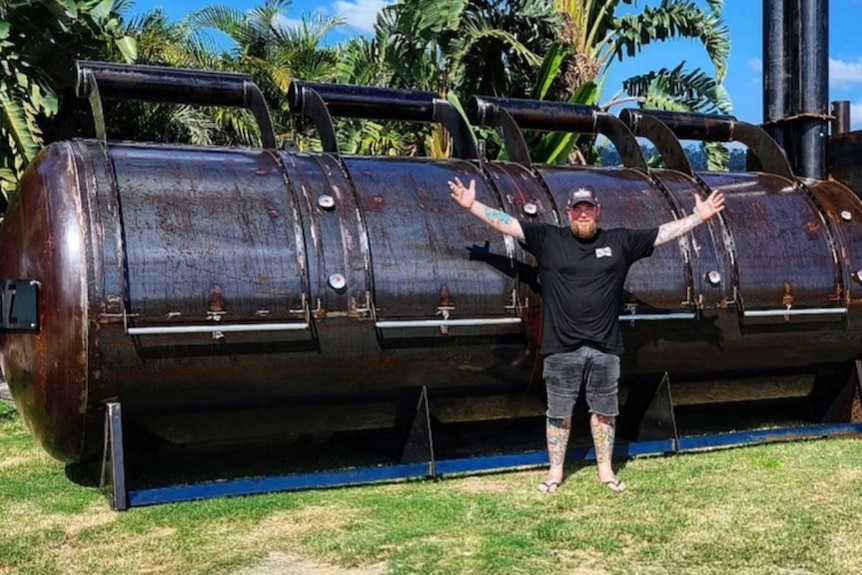 A man wearing black waves his arms in front of a giant brown meat smoker
