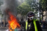 A man in a mask and a yellow vest in the foreground with a bright orange fire.