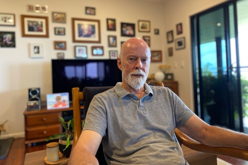 A man, bald with a gray beard, sits in a living room, blurry behind him is a wall of family photos. 