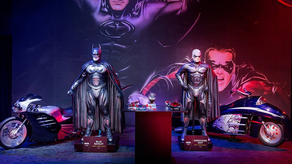 Two muscular Batman and Robin costumes displayed on a moodily lit stage