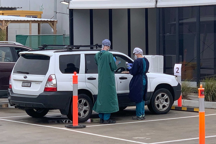 Two people in medical gowns and caps and gloves stand by a while car parked at a testing site in Altona North.