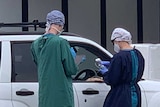 Two people in medical gowns and caps and gloves stand by a while car parked at a testing site in Altona North.