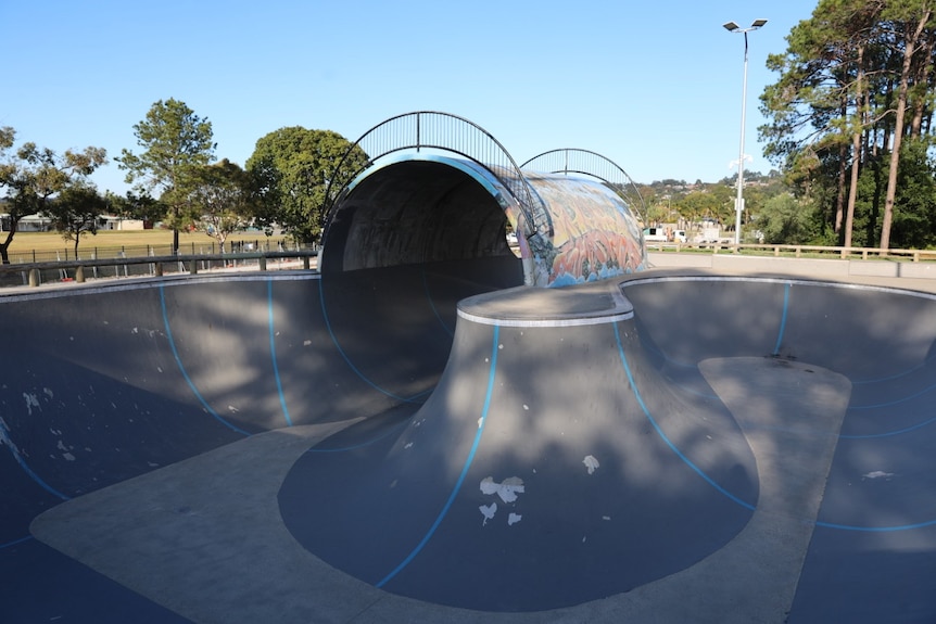 Grey concrete skate park with bright graffiti tunnel, green trees and blue sky in background