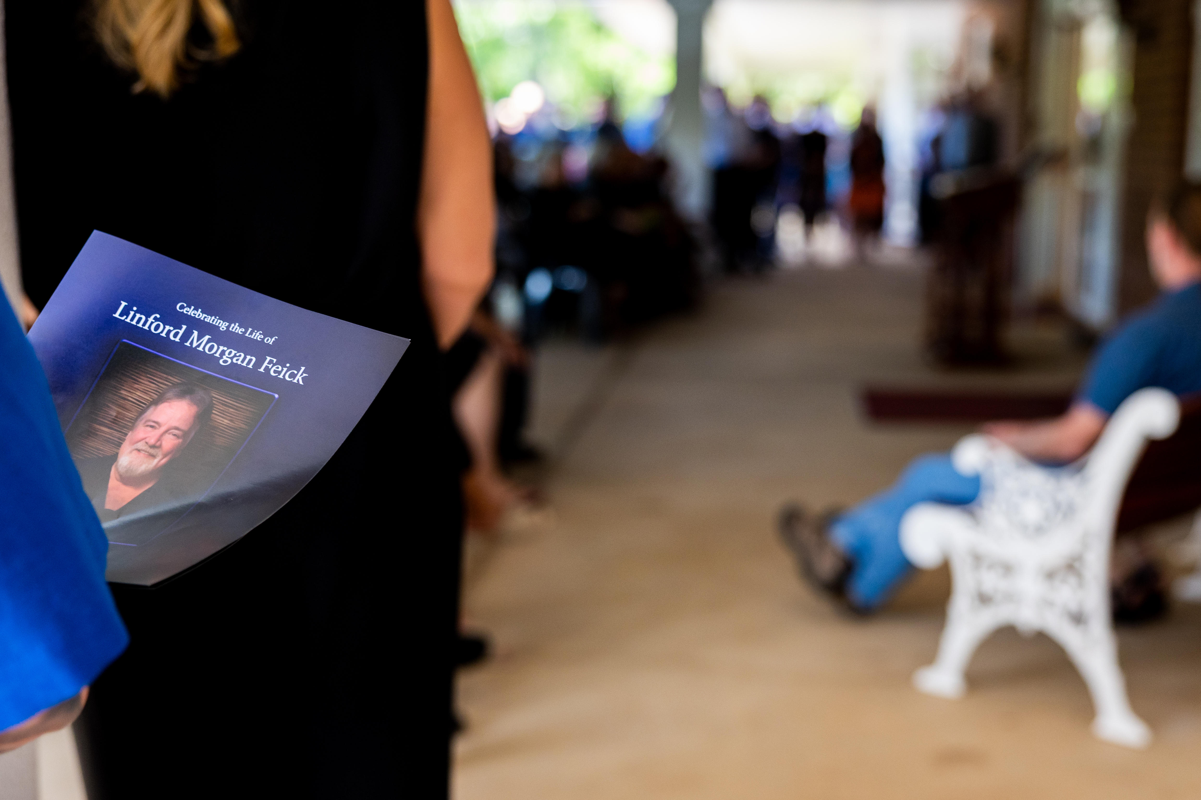 A blurred background of sombre people sitting, with a funeral program booklet in focus which reads 'Linford Morgan Feick'
