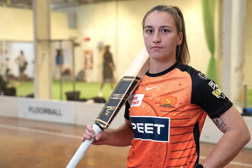 Mikayla Hinkley stands with a cricket bat propped on her shoulder.