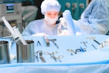 A surgery room, with a blurry figure of a female doctor operating and a nurse partially obscured, with instruments in foreground