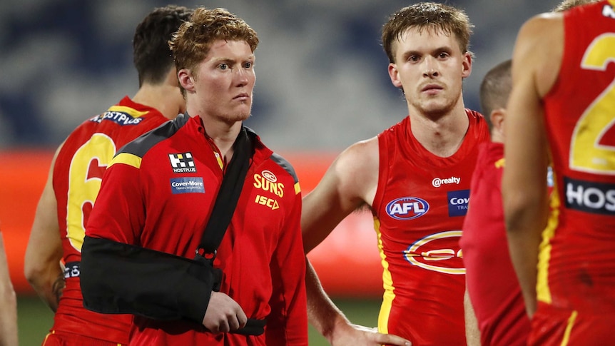 A young AFL player with his arm in a sling stands next to a teammate after a game.