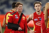 A young AFL player with his arm in a sling stands next to a teammate after a game.