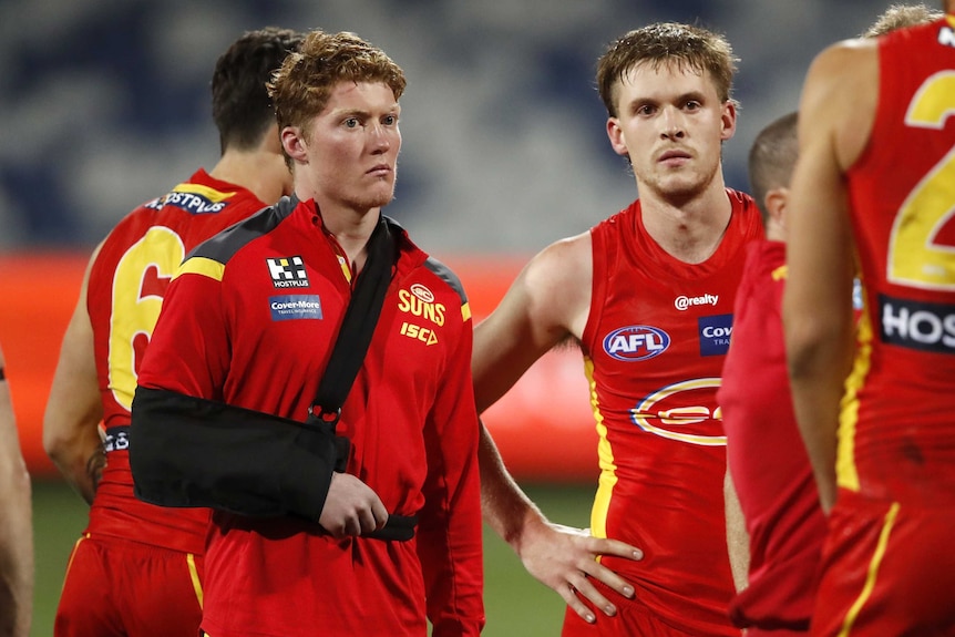 A young AFL player with his arm in a sling stands next to a teammate after a game.