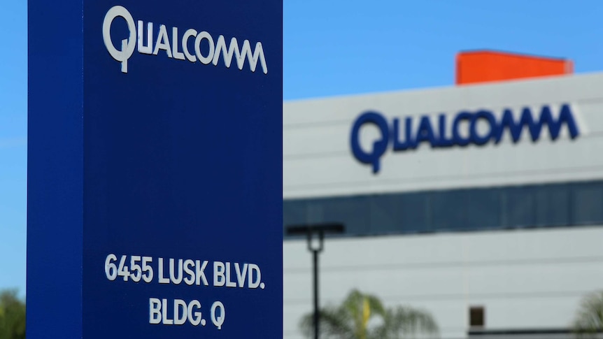 A blue sign with the words Qualcomm and an address is seen in front of another grey building with Qualcomm written in blue.