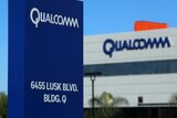 A blue sign with the words Qualcomm and an address is seen in front of another grey building with Qualcomm written in blue.