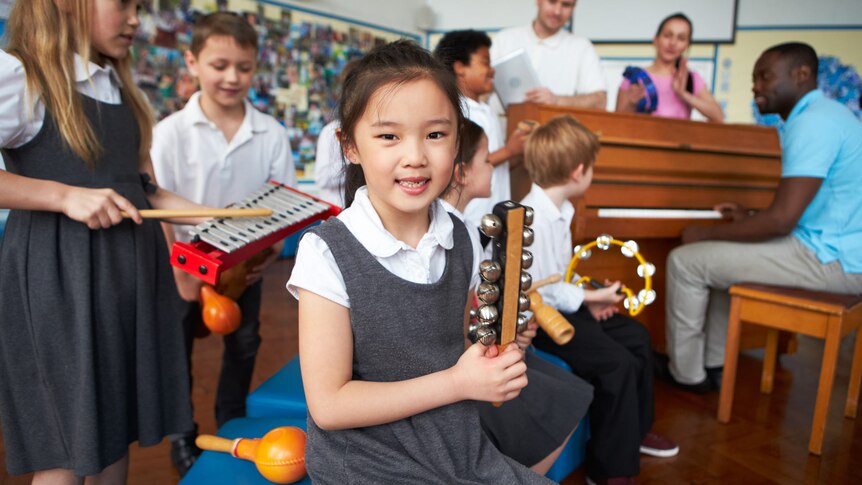 A young student in school uniform holds a jingle stick as her classmates play other percussive instruments behind her.